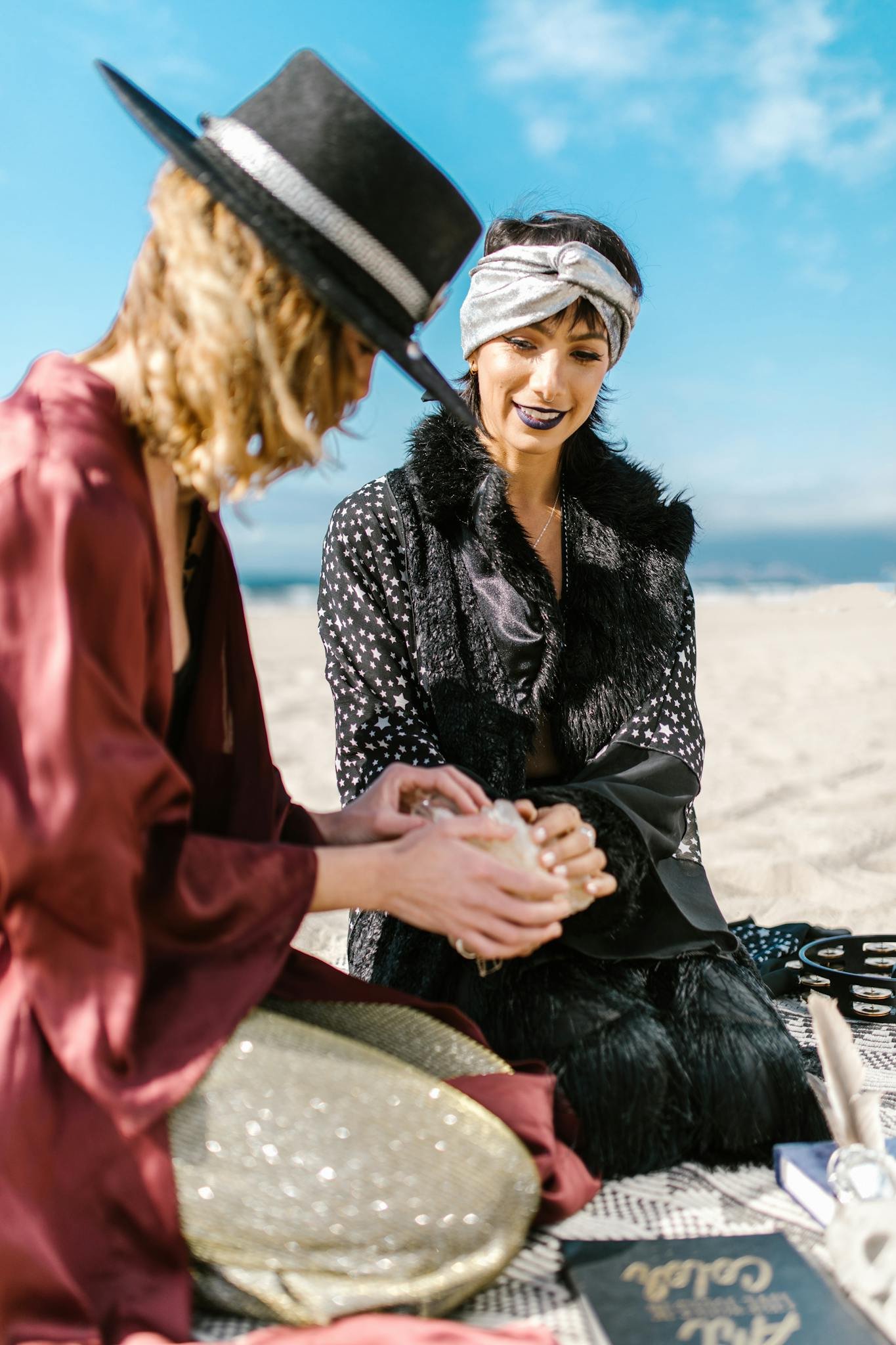 Two women engage in a spiritual reading on a sunny beach, embracing bohemian fashion and mysticism.